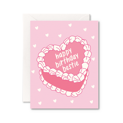 The Pretty by Her Happy Birthday Bestie Card is a pink A2 greeting card with a heart-shaped cake illustration, white frosting text, and small white hearts. Made in Canada from high-quality cardstock. An envelope peeks out behind the card.