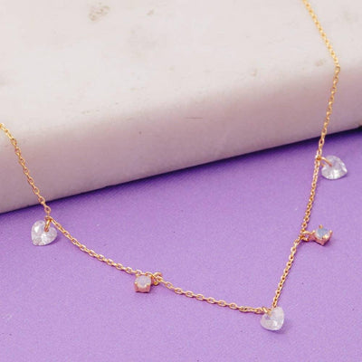 The Fame Accessories Gold Dipped Cz Heart Station Necklace, made in Korea, features five clear heart-shaped pendants and is displayed on a purple surface with a white marble slab in the background.
