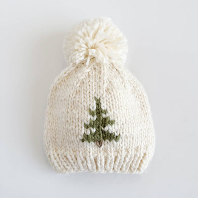 The Figgy Friends Tree Knit Baby Beanie is a cream-colored, hand-knit hat with a large pom-pom and small green pine tree on the front—perfect for fall and winter—shown on a white background.