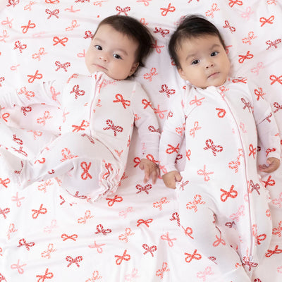 Two babies lie on a bed, holding hands and gazing at the camera in matching Angel Dear Candy Cane Bows Ruffle Back Two-Way Zipper Footies. The white pajamas feature red and pink bow patterns, echoed by the bow design on the bed sheet.