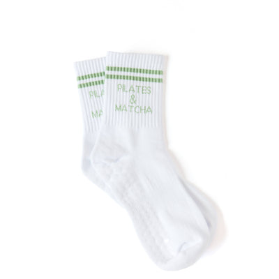 The Pilates University 1883 “Pilates & Matcha - (White)” crew socks feature white ribbed fabric, green stripes, and “PILATES & MATCHA” in green on the sides. Socks are shown laid flat on a white background.