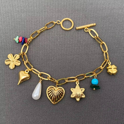 The Este Charm Bracelet by Mio Queena is an 18K gold plated, non-tarnish stainless steel accessory with clover, heart, pearl drop, star, turquoise bead, and colorful stone charms—shown on a gray textured surface.