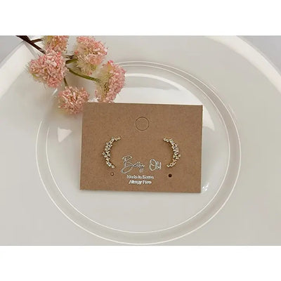 The BETTYOH INC CZ Moon Shaped Post Earrings are shown on a brown card atop a white plate, with pink flowers in the background. They feature delicate gold tones, small white beads, and silver posts.