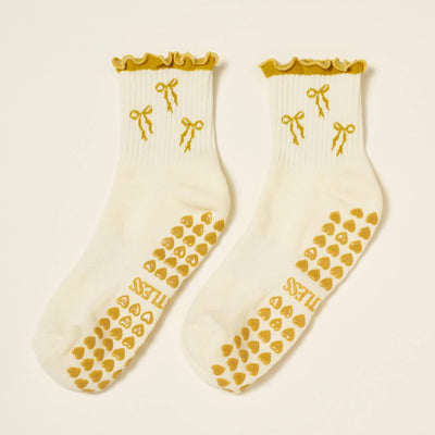 Effortless Clubs Bow Ruffle Grip Sock [Gold] is a white womens sock with yellow ruffled cuffs, bows, and non-slip gold heart grips. TILASS is printed on the sole. Made from soft fabric, perfect for Pilates.