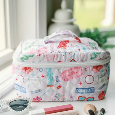 The Gracefully Made Taylor Swift Make Up Toiletry Bag, featuring colorful illustrations of shoes, dresses, and street signs, sits on a white surface. Nearby lie white round-framed sunglasses, a makeup brush, and red lipstick against a serene blurred green outdoor backdrop.