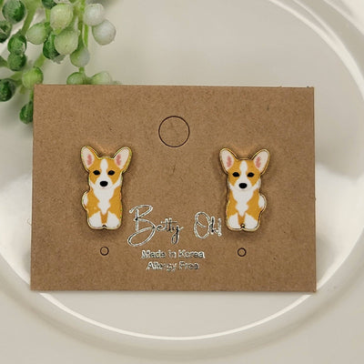 Corgi Enamel Post Earrings by BETTYOH INC, shaped like a brown and white corgi, are displayed on a brown card with Made in Korea and Lead Nickel Free, set on a white plate with a green faux plant nearby.