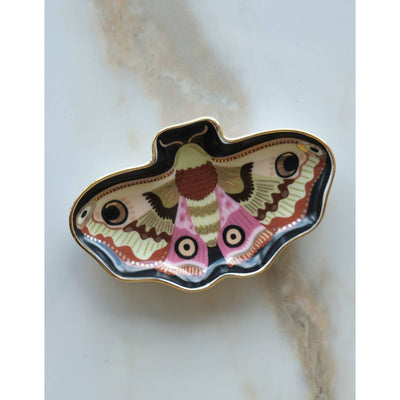 The Moon Moth Porcelain Trinket Dish by Idlewild Co. is a giftable ceramic dish shaped like a colorful moth, featuring pink, black, yellow, and green patterns with elegant 10k gold edges, displayed on a marble surface.