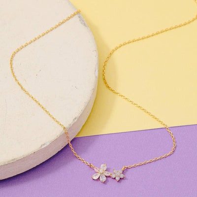 The Athens Pendant Necklace by Fame Accessories features a gold-dipped brass chain with a small floral pendant embellished with clear gemstones, shown on a round white surface against pastel yellow and purple hues.