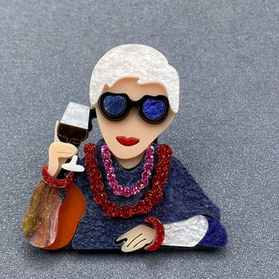 The Mio Queena Iris Acrylic Brooch features a woman with short white hair, blue heart sunglasses, red glittery jewelry, and a wine glass on a gray background—an eye-catching accessory and unique gift for jewelry lovers.