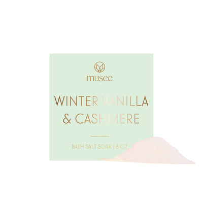 A pale green Musee Winter Vanilla & Cashmere Mini Bath Salt Soak packet (6 oz) sits on a white background beside a small pile of fine, white bath salts.
