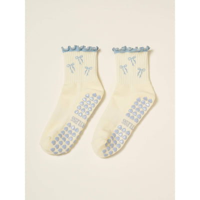 Effortless Club’s Bow Ruffle Grip Sock [Dusty Blue] features cream fabric with blue ruffled cuffs, small blue bow designs, and blue non-slip grips. "TILLES" is printed on one cushioned footbed.