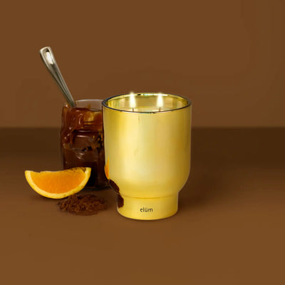 A shiny gold tumbler sits beside a glass jar of chocolate spread with a spoon, a slice of orange, Elum Designs Home’s Pumpkin Creme Brulée 21 oz Maeve Glass Candle, and coffee grounds, all set against a brown background.