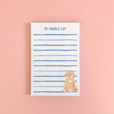 The Gert & Co to doodle notepad features blue-lined pages with a cute watercolor brown Goldendoodle in the bottom right corner on a light pink background.