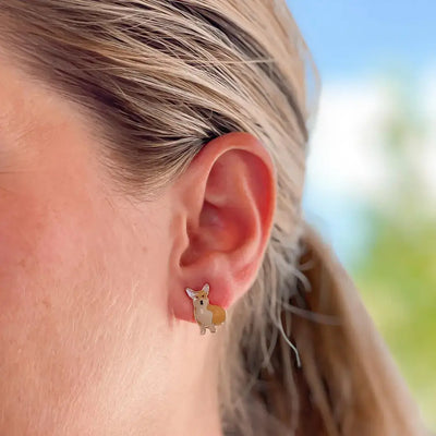 A close-up shows a person wearing Prep Obsessed Wholesales Corgi Enamel Stud Earrings, nickel free and shaped like a cute brown and white dog, against a blurred blue sky and greenery—an ideal accessory for any dog lover.