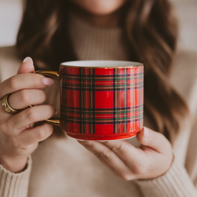 A person wearing a beige sweater holds the Sweet Water Decor Red Plaid Coffee Mug. Only their hands and a section of their long, wavy brown hair are visible.