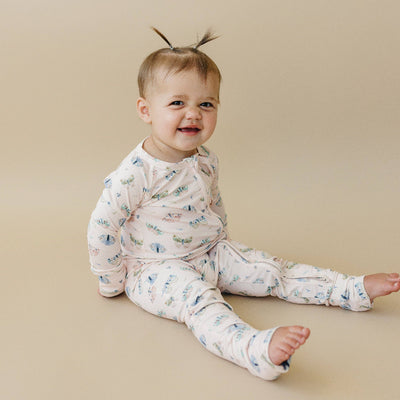 A smiling baby with light hair in pigtails wears the Little Joy Co. Blushing Butterfly Bamboo Zippy Romper, sitting on a beige background.