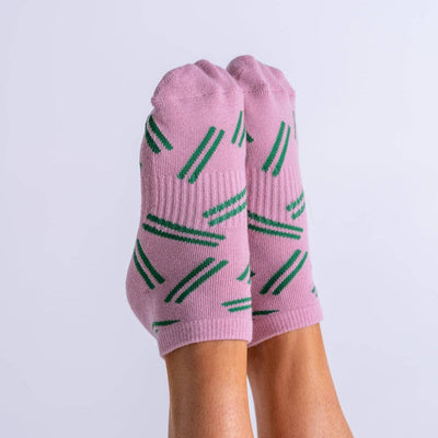 A pair of feet wear light pink Slater Stripe Pilates Ankle Grip Socks with green diagonal stripes by Reformer Loops, shown against a plain white background.