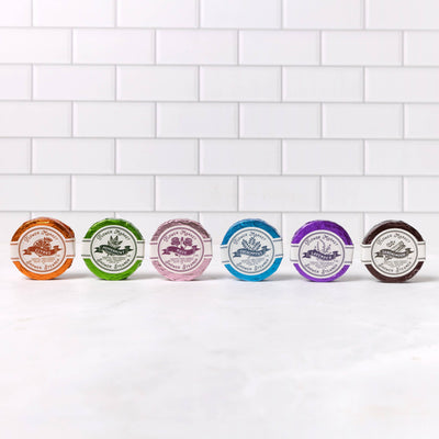 Six Aromatherapy Shower Steamers from 123 Farm in various colors are arranged in a row on white marble, bringing spa-like calm, with a white subway tile wall in the background.