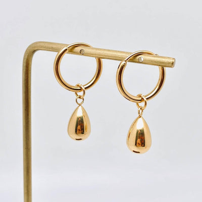 The Mio Queena Teardrop 18k Gold Plated Huggie Earrings are shown on a minimalist gold stand against a plain white background.