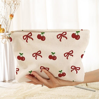 A hand holds the Judson & Company Corduroy Bows & Cherries Pouch, a cream item adorned with red cherry and ribbon patterns, topped with a zip closure. Behind it, dried flowers in a vase sit elegantly on a table beside a white curtain.