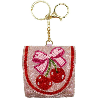 The Bow Cherries Seed Beaded Air Pod Case & Key Ring by Judson & Company features two red cherries with a pink bow. Perfect Air Pod size, it includes a gold keyring and chain for stylish, easy access.