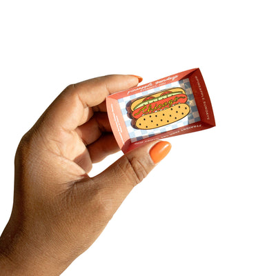A hand with orange nails holds a small red box from Pineapple Sundays Design Studio, showcasing the Chicago Style Hot Dog Pin—a colorful enamel pin with "Chicago" in yellow on the illustrated hot dog.