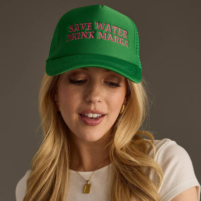 A person with long blonde hair is wearing a white top and a Save Water Drink Margs Trucker Hat by Panache Apparel Co. The green cap with pink lettering features an adjustable snapback closure, adding a casual vibe as they look down, smiling slightly against the gray backdrop.