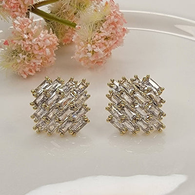 Fancy CZ Post Earrings by BETTYOH INC feature a square design in gold and silver, accented with a crisscross pattern and sparkling clear stones. Softly blurred pink flowers sit in the background on a white surface.