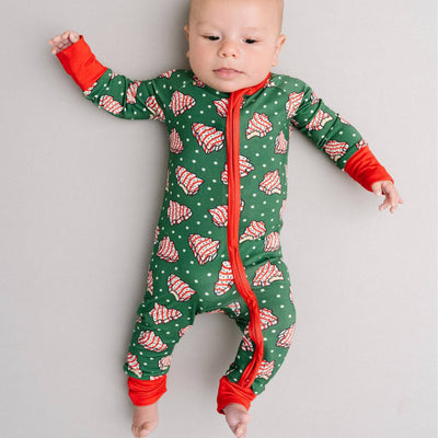 A baby lies on a light surface wearing the Little One Shop Tree Treats Bamboo Sleeper, featuring green fabric with red and white Christmas tree patterns, fold-over mittens, and red cuffs.
