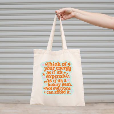 A hand holds the Sonny Rising Think of Your Energy As Expensive Canvas Tote Bag, featuring colorful retro text on beige canvas. A metal shutter serves as the background.