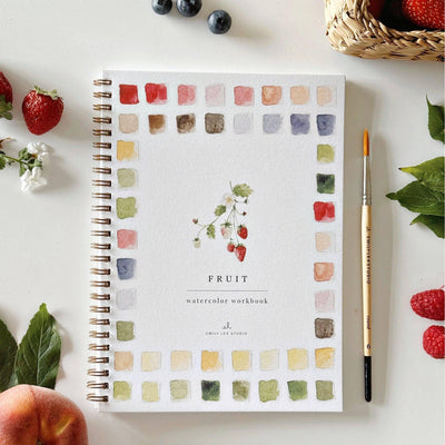 The Fruit Watercolor Workbook by emily lex studio, featuring a fruit cover and color swatches, rests on a white surface with strawberries, raspberries, green leaves, a paintbrush, and a strawberry basket—an ideal gift for artists.