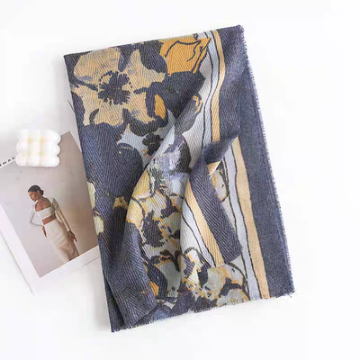 The Niello Fall Print Scarf by Pretty Persuasions, featuring a blue, yellow, and grey floral pattern in 100% polyester, is displayed on a white surface beside a wedding photo and a white bubble-shaped candle.