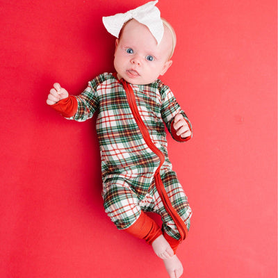A baby wearing the Little One Shop Christmas Plaid Bamboo Sleeper with fold-over mittens lies on a solid red background, gazing up with wide blue eyes and accessorized with a white bow headband.