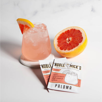 A glass of pink Paloma cocktail with ice sits beside a sliced grapefruit and two packets of NOBLE MICKS Paloma Single Serve Craft Cocktail Mix made from natural ingredients. Garnished with a grapefruit wedge, the drink rests elegantly on a white marble background.