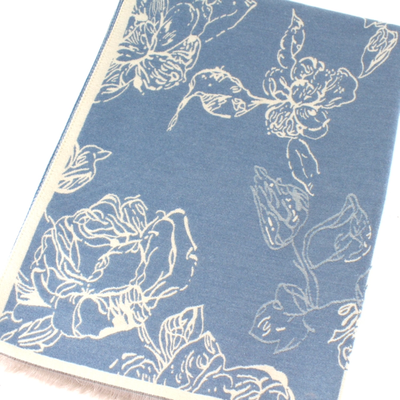 The Lorelai Reversible Scarf by Pretty Persuasions features blue polyester-viscose fabric with a white floral outline of large flowers and leaves, bordered edges, and short fringe on one end.