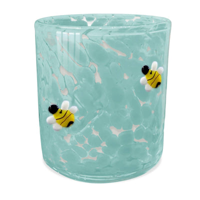 The SOi Company presents the Bees Knees 12oz Blue Bees Confetti Candle, featuring a light blue, speckled glass tumbler with textured honeycomb-inspired appearances and adorned with yellow-and-black striped bees with white wings.