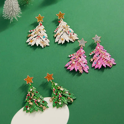Three pairs of Golden Stella Decked Out Christmas Tree Earrings—white, pink, and green—each with colorful ornaments and a glittery star, displayed on a green surface. Hypoallergenic titanium posts offer comfortable wear.