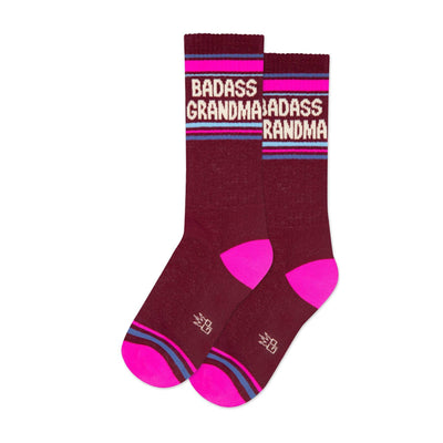 The Badass Grandma Gym Crew Socks by Gumball Poodle are maroon cotton mid-calf socks with pink heels and toes, blue and pink stripes, and BADASS GRANDMA in bold white and pink lettering—perfect for any proud grandma.