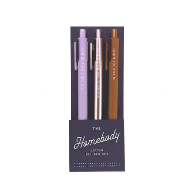 The Homebody Jotter Gel Pen Set by Ruff House Print Shop includes three vibrant gel pens—purple, rose gold, and brown—with unique vertical phrases, all in a dark blue holder labeled "Homebody Gel Pen Set.