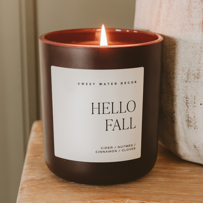 The Sweet Water Decor Hello Fall 15 oz Candle in a matte jar features notes of nutmeg, cinnamon, and cloves—perfect cozy fall decor for any wooden surface.