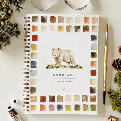 The Woodland watercolor workbook by emily lex studio features a bear cover with a swatch border. Spiral-bound and perfect for gifting, it includes a paintbrush; dried plants, ribbon, and a paint tube are shown arranged nearby.