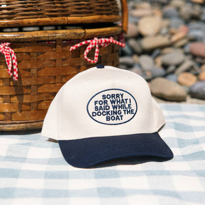 The Ruby Moon Sorry For What I Said While Docking The Boat Baseball Hat in white and navy blue sits on a checkered blanket, its playful message visible against a backdrop of smooth stones.