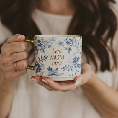 A person in a white shirt, wearing a simple gold ring, holds the Sweet Water Decors Best Mom Ever Coffee Mug. This Fine Bone China mug showcases a Blue Delft Design and best MOM ever inscribed in gold, complemented by its gleaming golden handle.