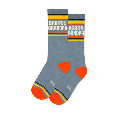 The Gumball Poodle Badass Grandpa Gym Crew Socks feature blue fabric, orange toes and heels, yellow, white, and black stripes, plus “BADASS GRANDPA” in bold white letters near the top. Made in the USA for comfort and style.