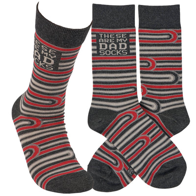 Three cotton socks with a gray base, featuring striped patterns in gray, white, and red. They display the text THESE ARE MY DAD SOCKS from the brand Primitives by Kathys product line: These Are My Dad Socks.