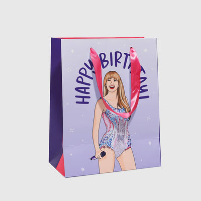 The Brittany Paige Happy BirthTAY Gift Bag is light purple with pink handles, showcasing a woman in a sparkly outfit holding a microphone with Happy BirthTAY written above, perfect for delighting Swifties.