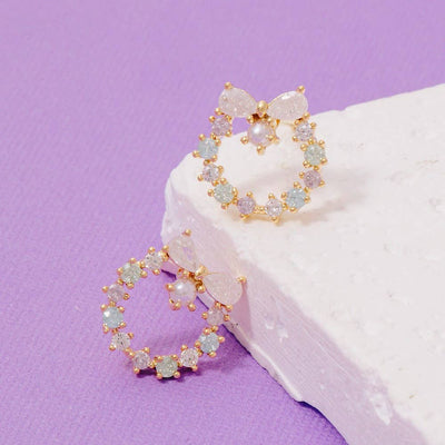Fame Accessories’ Gold Dipped Cz Tiara Circle Earrings, featuring sparkling gemstone accents and bow designs, are displayed on a light textured white block against a purple background.