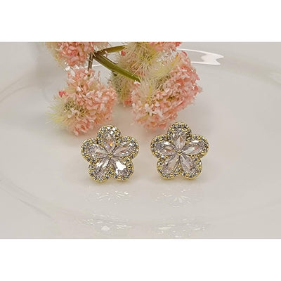 The Faye CZ Flower Post Earrings by BETTYOH INC feature gold flower shapes with clear cubic zirconia, shown on a white surface with pink flowers. Elegant and sparkling, they’re perfect for elevating any outfit.