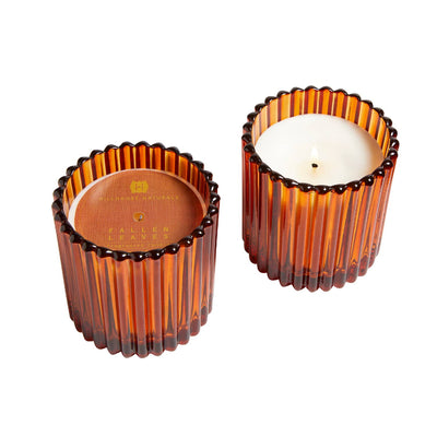 Two amber ribbed glass containers from Hillhouse Naturals/Field+Fleur; one is a Fallen Leaves Small Glass Candle (7oz), the other has a wooden lid with gold logo. Both feature scalloped edges and a modern, elegant design.