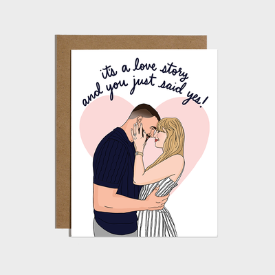 The "You Just Said Yes Engagement Card" by Brittany Paige is an A2 card with a couple in a pink heart and "It's a love story and you just said yes!" above, printed on quality paper with a matching envelope included.